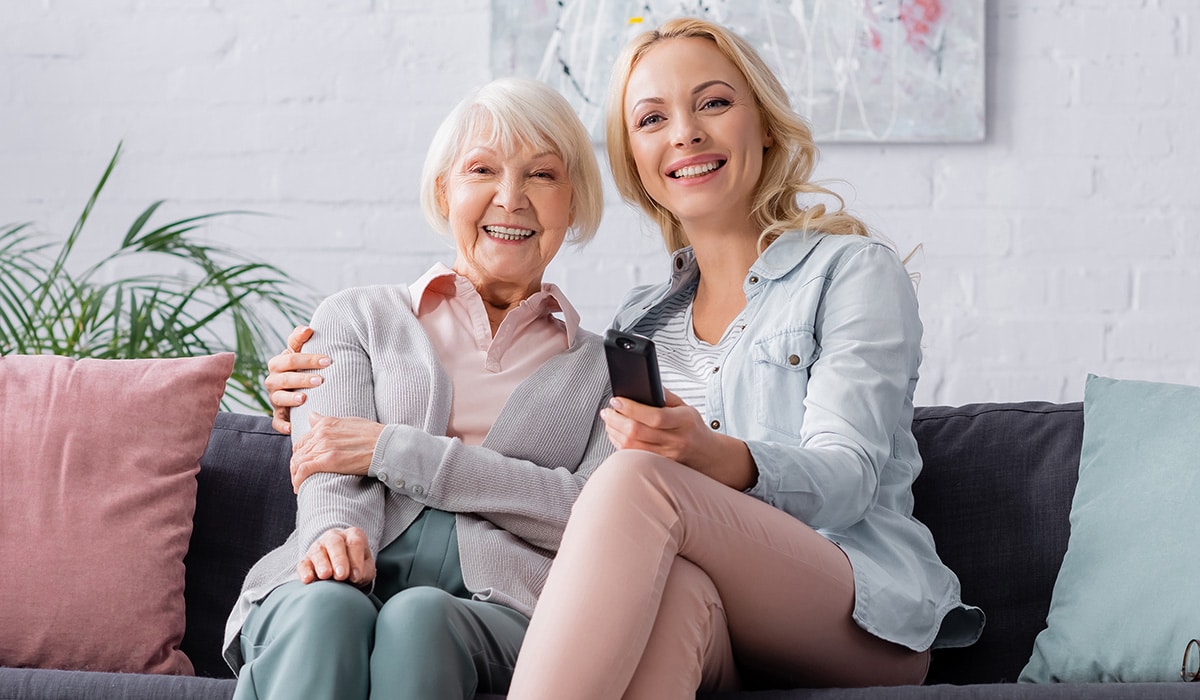 Cheerful woman and her elder mother, both smiling as they hold a remote control and watch TV.