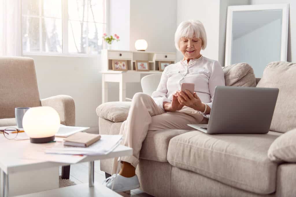 Independent senior woman texting on her phone with a laptop nearby and smiling happily while sitting on the couch.