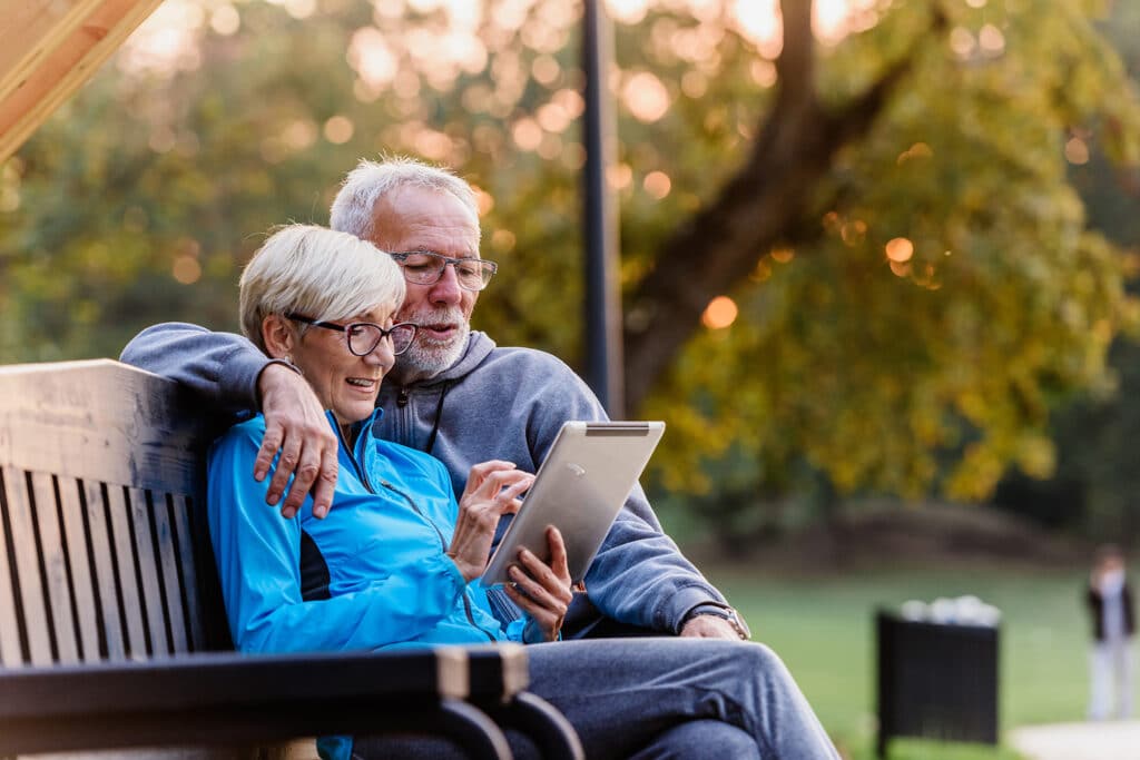 Senior couple sitting on a bench outside in a courtyard or park using a wifi tablet