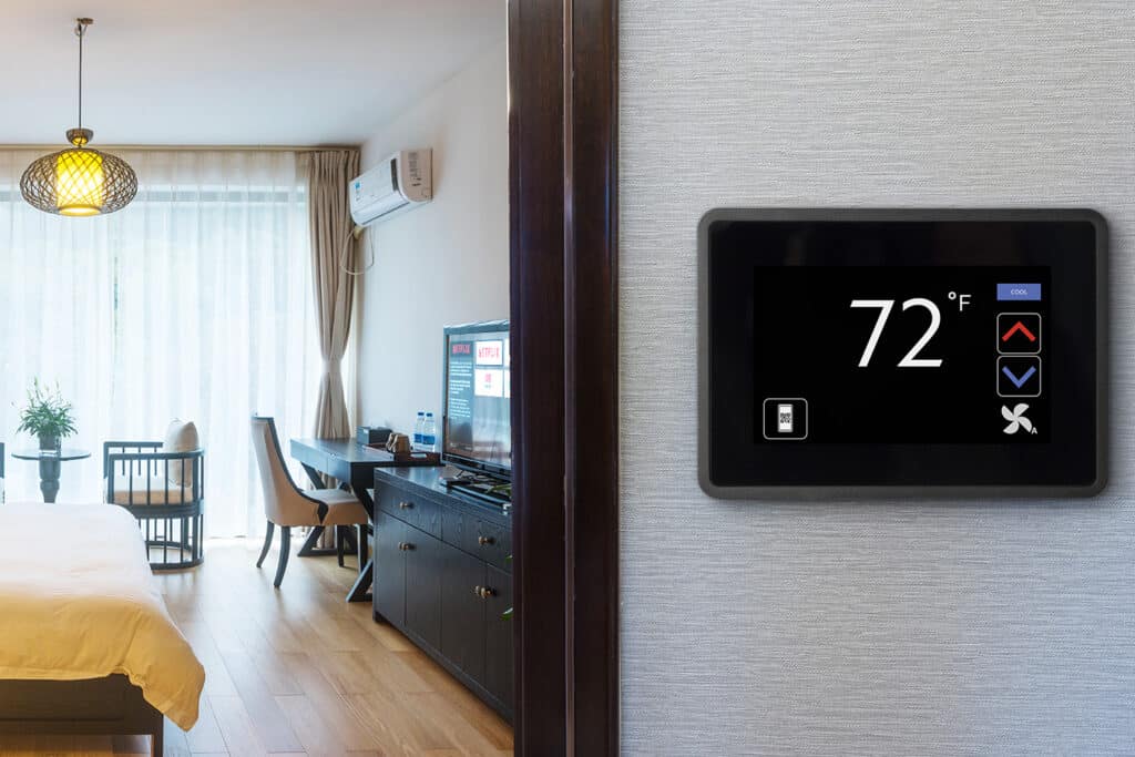 Modern, well-lit room with a smart thermostat displaying 72°F on the wall. There's a bed, desk, TV, air conditioner, and elegant pendant light.