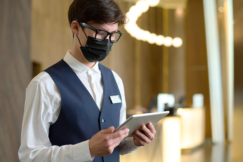 A person wearing a black mask, vest, white shirt, and glasses is focused on a tablet inside a well-lit, modern-looking interior space.