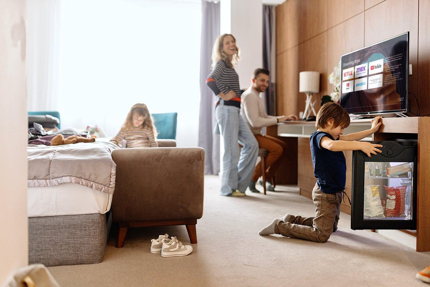 This image shows two children and two adults in a hotel room. One child opens a mini-fridge, a person works at a desk, and both overlook the kids.
