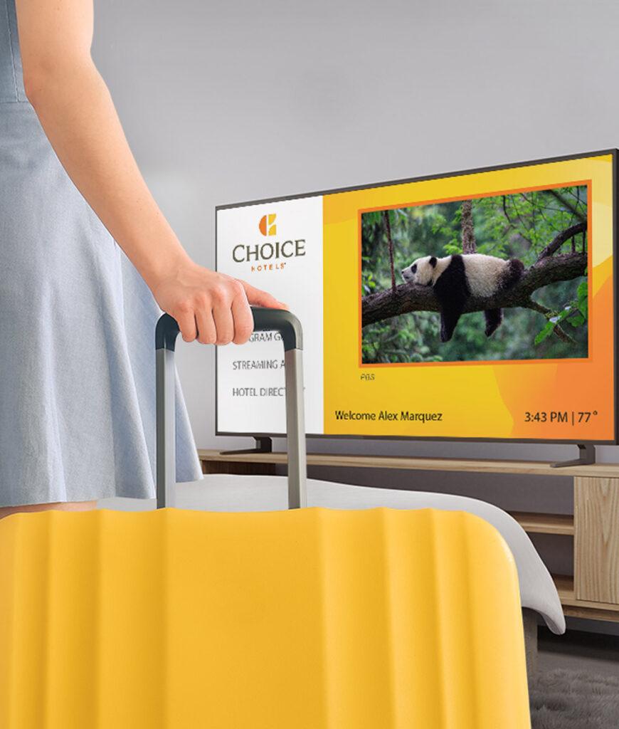 A person in a blue dress is standing next to a bed with a yellow cover, holding a suitcase handle, with a TV screen displaying a panda.