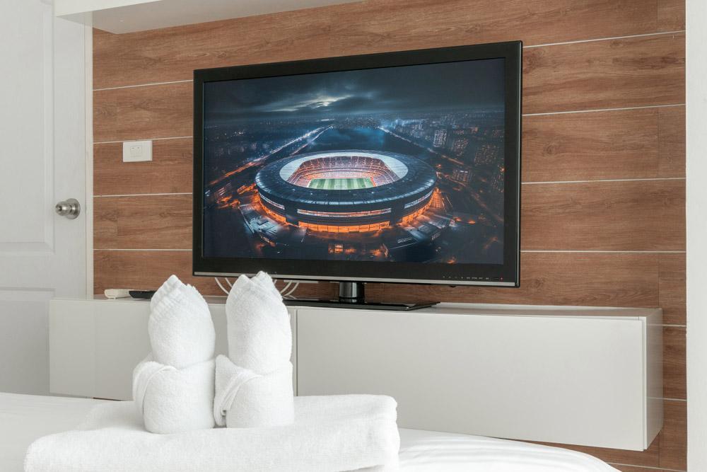 A modern bedroom with a flat-screen TV displaying an illuminated stadium at night. There are neat white towels shaped like bunnies on the bed.