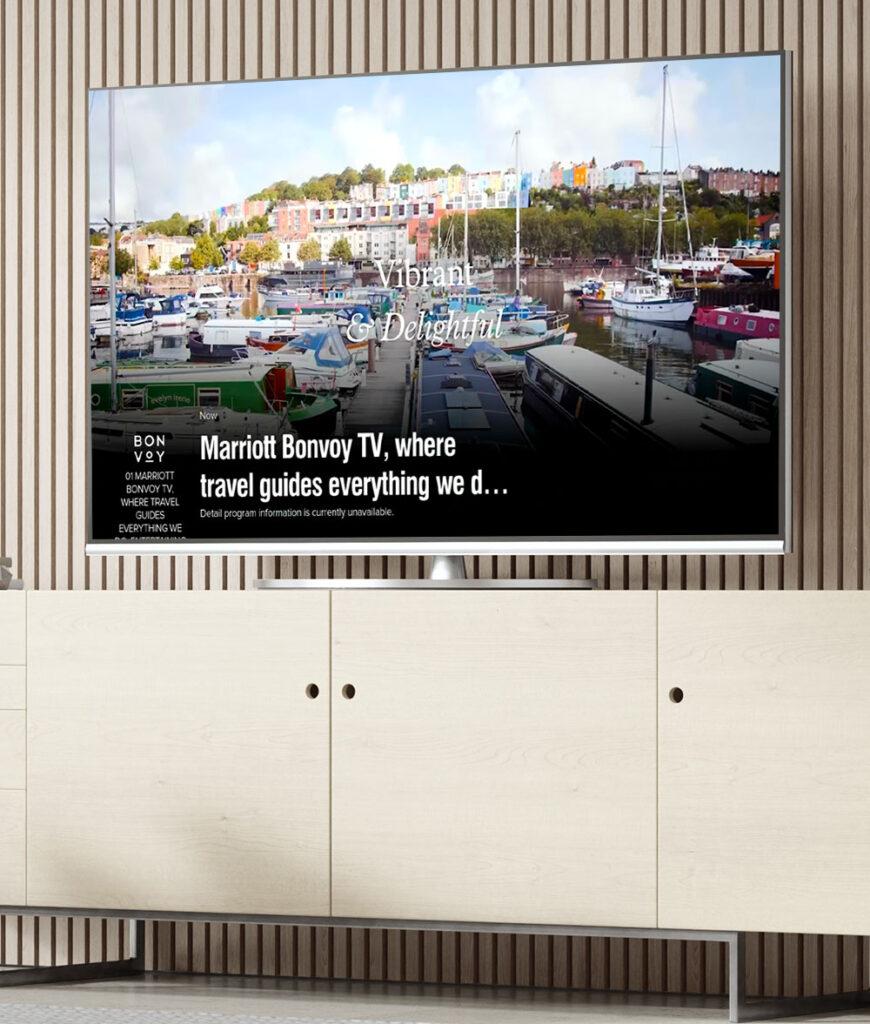 Modern interior with a large wall-mounted TV displaying a harbor scene with boats and a promotional message for Marriott Bonvoy TV. Below is a wooden cabinet.