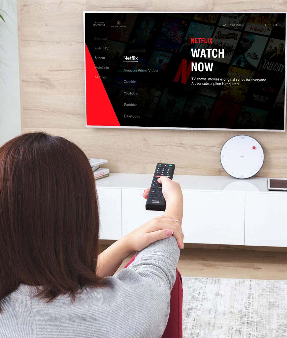 A person sitting on the floor points a remote at a TV displaying a Netflix screen with various streaming options. A clock hangs beside the TV.