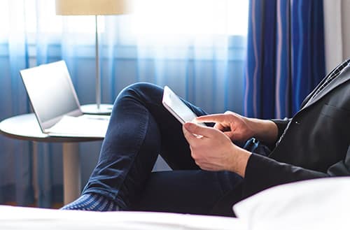 A person reading on a tablet in a hotel room with a laptop nearby.