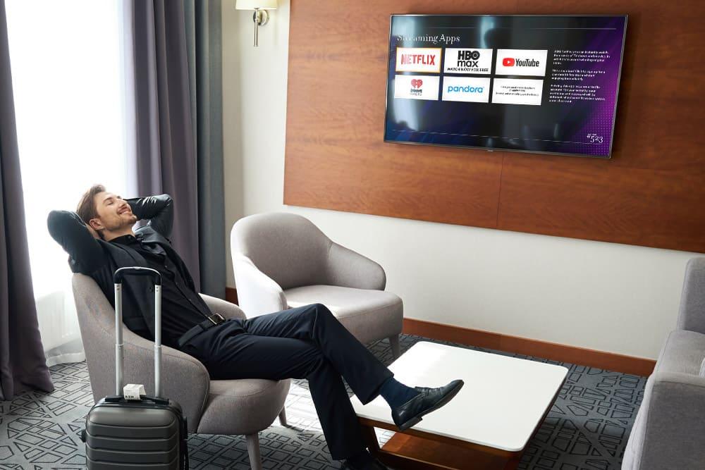 Man reclining in a chair in a hotel room with the TV on displaying Enseo's Streaming Apps screen